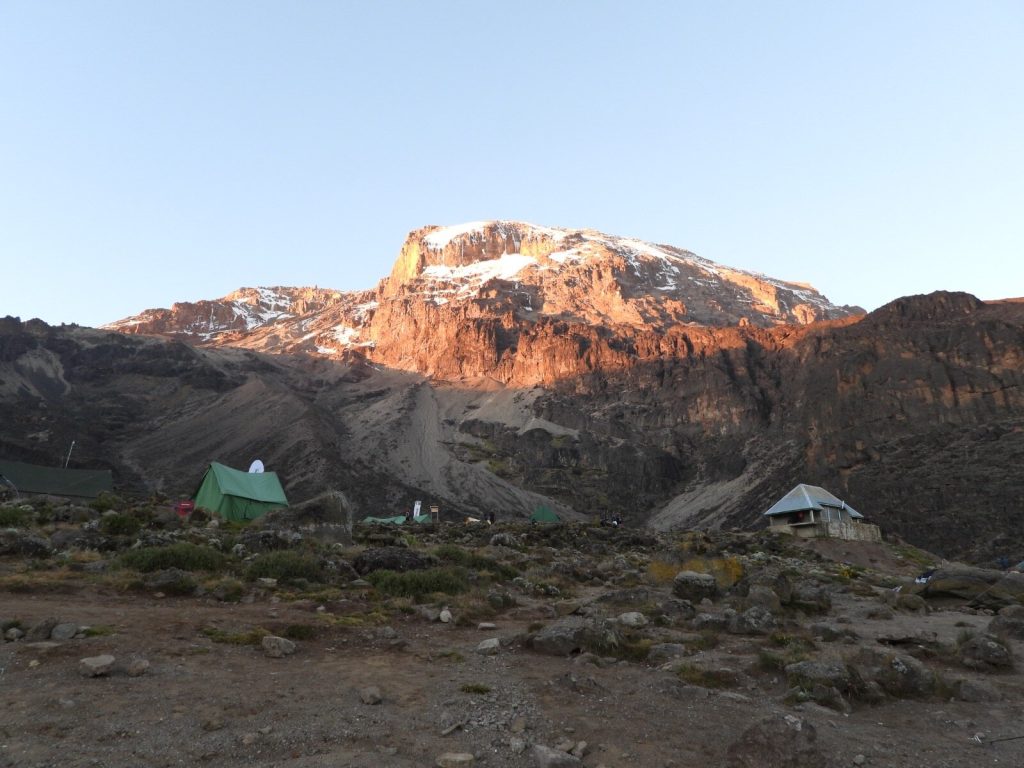 Green camp tents and hut below sunlit cliffs at dawn on Mount Kilimanjaro