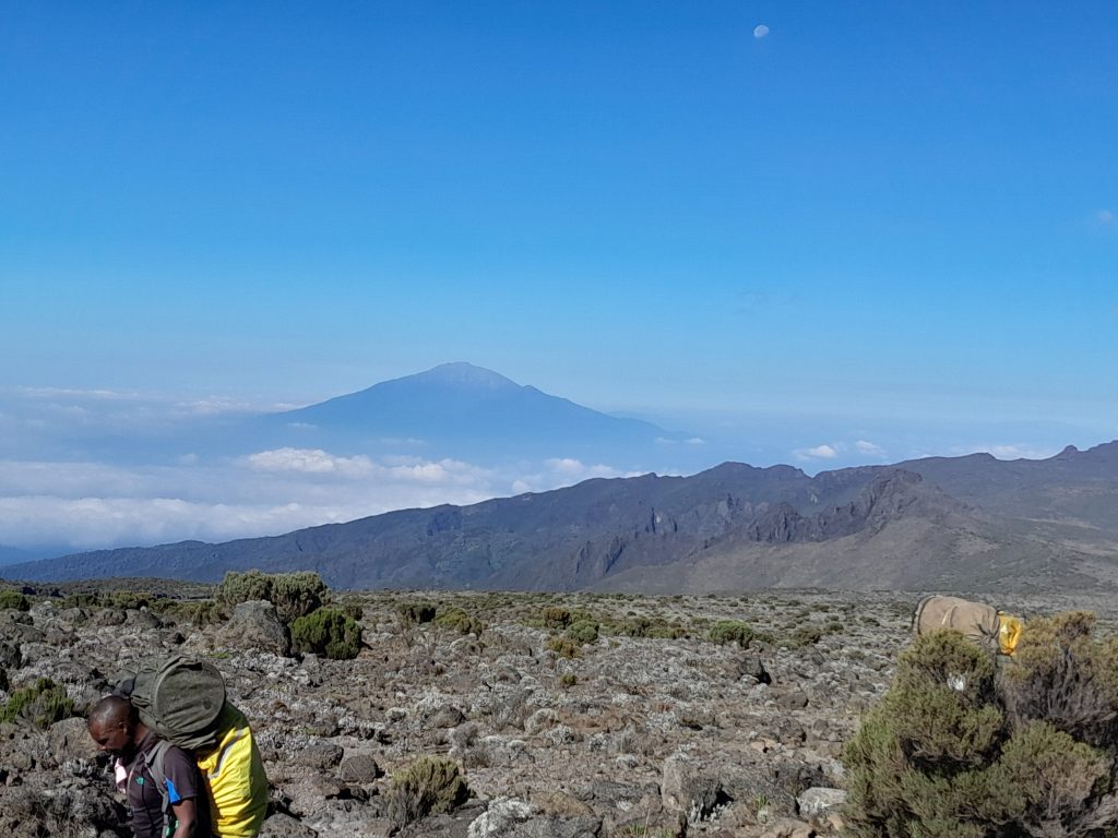 Porter carrying a trekking duffel on Kilimanjaro with a distant volcanic cone behind
