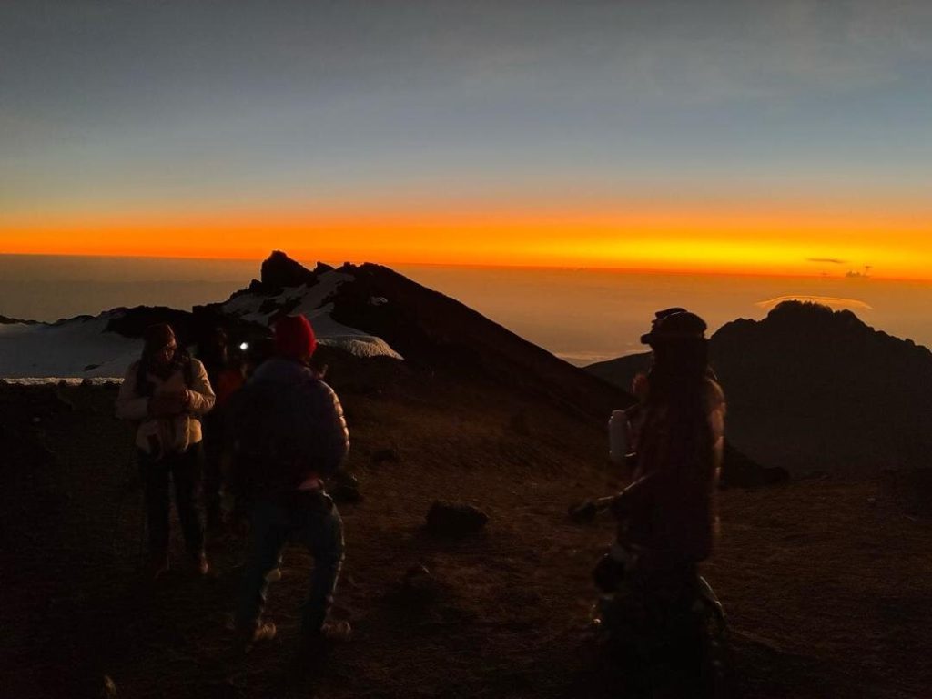 Trekkers silhouetted on a ridge under an orange dawn sky on Mount Kilimanjaro