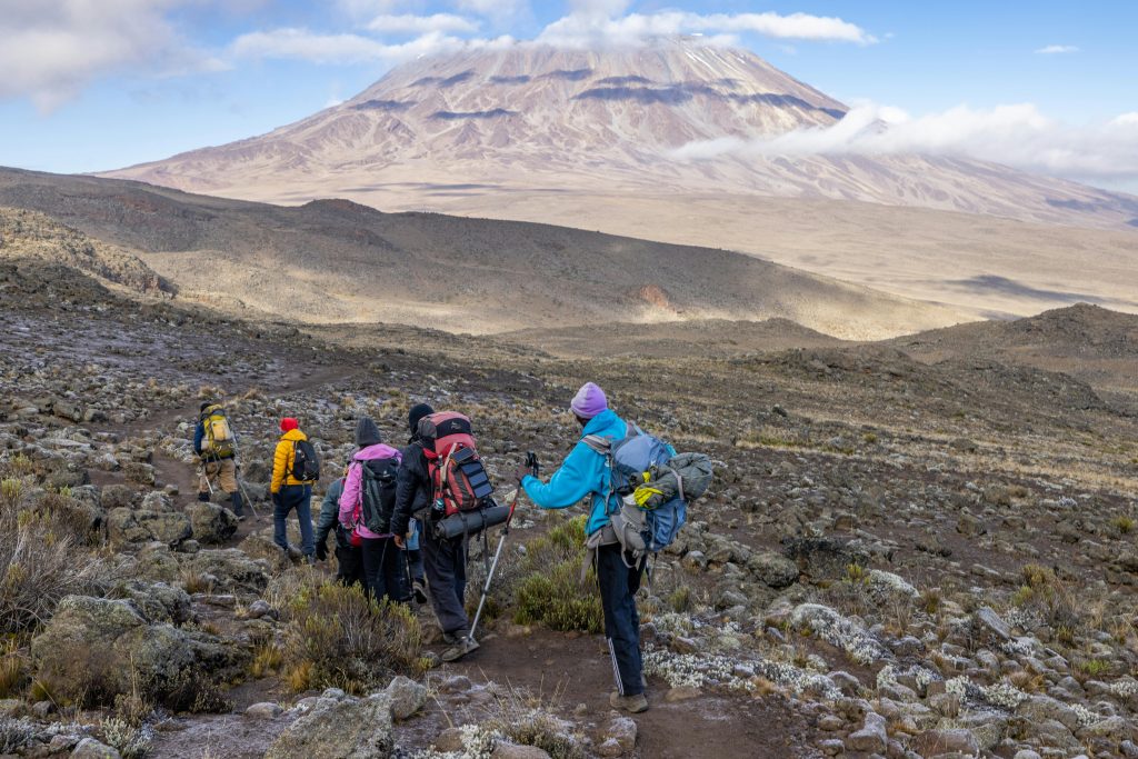 Six trekkers walking a scree trail towards the bare summit dome of Mount Kilimanjaro