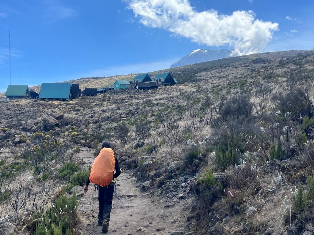 Trekker with orange pack below green roofed huts on Mount Kilimanjaro