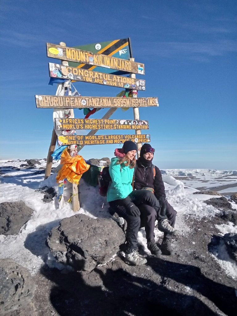 Two trekkers seated at the sunlit uhuru peak signboard on Mount Kilimanjaro